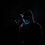 Silhouette of a person in a hoodie wearing a glowing LED mask against a dark background.